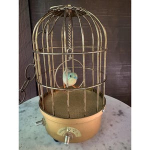 May include: A gold-toned metal birdcage lamp featuring a small, light blue artificial bird perched on a circular swing inside. The base is a solid gold cylinder with a mesh floor, two metal control knobs, and text reading INTERVAL ON OFF.