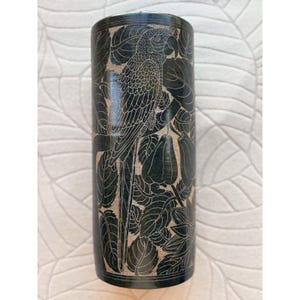 May include: A cylindrical, dark-colored vase with a carved design featuring a parrot perched among leaves and flowers. The parrot and foliage are in a lighter color, creating a contrast against the dark background. The vase is a decorative item.