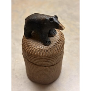 Puede incluir: Un recipiente cilíndrico de cerámica con un exterior texturizado de color tostado que presenta estrías horizontales. Una pequeña figura de oso de color marrón oscuro con el hocico más claro está fijada en la tapa, sobre una superficie tallada.