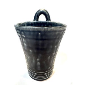 May include: A dark blue ceramic vase with a textured surface and a loop handle. The vase has a wide mouth and a narrow base.