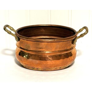 May include: A copper bowl with two brass handles. The bowl has a hammered texture and a rounded shape. The interior is a darker copper color, and the handles are a lighter gold color. The bowl is likely used for cooking or serving.
