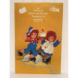 May include: A rectangular yellow cardboard packaging for a Hallmark Raggedy Ann and Andy Freewheeling Fun centerpiece. The graphic shows the two characters on roller skates, with Raggedy Andy holding a cake. It states 11 1/2 inches tall.