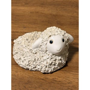 May include: A ceramic sheep figurine with a textured, off-white wool-like body. The sheep has a smooth, white face with black eyes and a small, smiling mouth. The ears are also white. The figurine is sitting on a wooden surface.