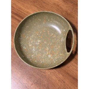 May include: A speckled green serving bowl with a handle. The bowl has a round shape and a speckled pattern of green, orange, and brown. The bowl is sitting on a wooden surface.