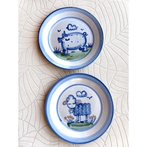 May include: Two ceramic plates with blue rims and hand-painted animal designs. One plate features a pig, the other a sheep, both in blue. The plates have a white background with blue accents and green grass details.