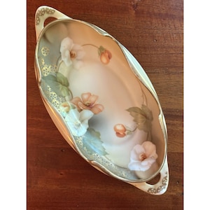 May include: An oval porcelain serving dish with two handles, featuring a cream-colored base decorated with hand-painted white and peach-colored flowers, green leaves, and gold filigree accents along the scalloped edges and handle areas.