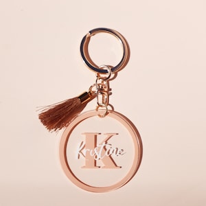 May include: A peach-coloured acrylic keychain with a gold chain and tassel. The keychain features a large letter "K" with the name "Kristine" written in white script.