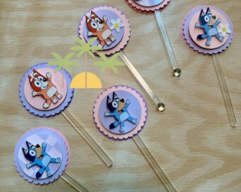 May include: Six circular cupcake toppers featuring Bluey and Bingo characters on layered scalloped cardstock in shades of pink and purple. Each topper is attached to a clear acrylic stick. One topper displays the blue text Bluey with a white flower.