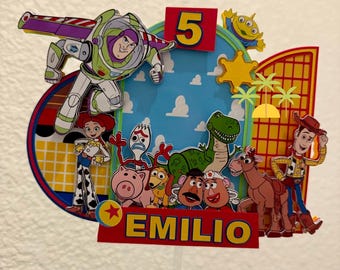Könnte beinhalten: Ein mehrschichtiger Tortenaufleger aus Papier mit Toy Story-Figuren wie Buzz Lightyear, Woody, Jessie, Rex und anderen. Das Design umfasst einen blauen Himmelshintergrund, eine rote Zahl 5 und ein rotes Banner mit dem Namen EMILIO in Gelb.