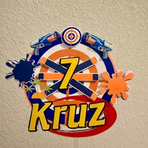 May include: A vibrant cake topper with a target, toy blasters, and the number 7. The name "Kruz" is in yellow with a black outline. Blue and orange splatter and palm tree graphics complete the design.
