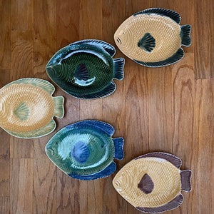 May include: Five ceramic fish-shaped plates in various shades of green, blue, and yellow. The plates are arranged in a semi-circle on a wooden surface.