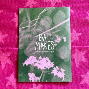 Puede incluir: Una tarjeta verde con el texto "BAT MAKES" en blanco, con un dise&ntilde;o floral. La tarjeta est&aacute; colocada sobre un fondo rosa con un patr&oacute;n de estrellas. Tambi&eacute;n se ve la direcci&oacute;n del sitio web.
