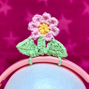 Könnte beinhalten: Rosa und gelbe gehäkelte Blume mit grünen Blättern, die an einem rosafarbenen Stirnband befestigt sind. Das Stirnband befindet sich auf einem weißen runden Objekt.