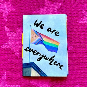 Könnte beinhalten: Ein kleines blaues Notizbuch mit dem Text "We are everywhere" in schwarzer Kursivschrift. Auf dem vorderen Umschlag ist eine Regenbogenflagge mit einem gelben Kreis oben links abgebildet.