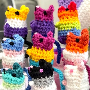 May include: A selection of colourful, handmade crochet cat figurines. The cats feature a variety of colour schemes, including rainbow, purple, yellow, and blue. Each cat has small black eyes and a tail. These whimsical cat toys are displayed on a clear shelf.