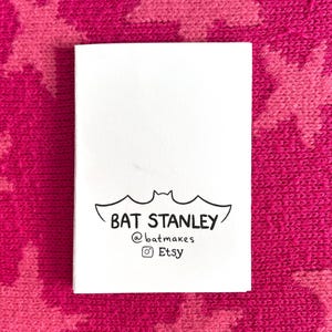 Peut inclure: Une carte blanche avec une illustration de chauve-souris noire et le texte "BAT STANLEY @batmakes Etsy" est pr&eacute;sent&eacute;e sur un fond rose avec un motif d'&eacute;toiles. La carte est rectangulaire et le texte est centr&eacute;.