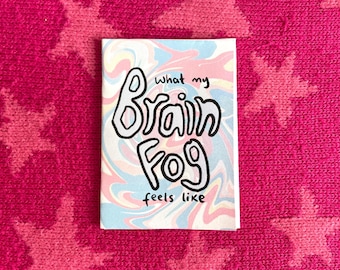 May include: A small, rectangular card with the words "what my Brain Fog feels like" in black, outlined bubble letters. The background features a swirling pattern of pastel pink, blue, and yellow colors.