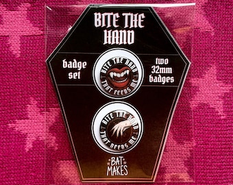 May include: A badge set featuring two 32mm badges with a vampire theme. One badge shows a mouth with fangs, the other a hand. Text reads "Bite the Hand That Feeds Me". The set is packaged in a clear plastic sleeve.