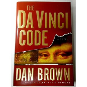 May include: A hardcover book titled The Da Vinci Code by Dan Brown with a red cover featuring gold lettering and a partial image of the Mona Lisa's eyes. The text A Novel and Author of Angels & Demons is printed on the front cover.