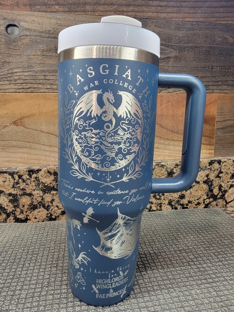May include: A blue stainless steel travel mug with a white lid and a black handle. The mug features a silver design of a dragon with wings and a quote that reads "There's nowhere in existence you could that I wouldn't find you. Violence" and "I have a thing for HIGHLORDS WINGLEADERS & FAE PRINCES".