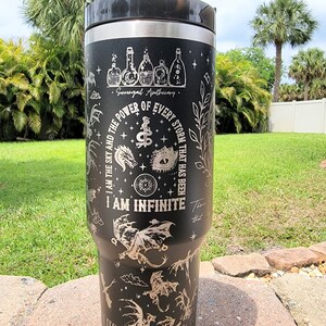 May include: Black stainless steel travel mug with a silver engraved design featuring dragons, stars, and the text "I am the sky and the power of every storm that has been. I am infinite. There that its".