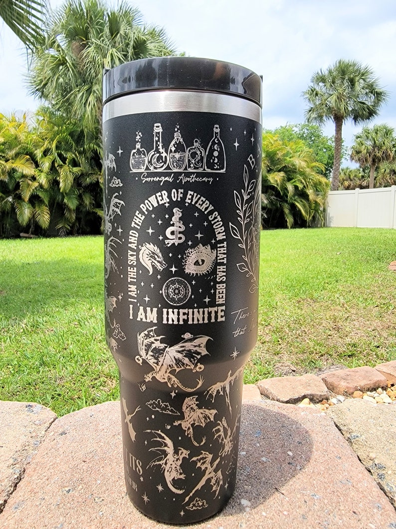 May include: Black stainless steel travel mug with a silver engraved design featuring dragons, stars, and the text "I am the sky and the power of every storm that has been. I am infinite. There that its".