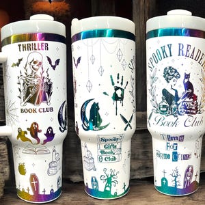 May include: Three white stainless steel tumblers with a rainbow iridescent finish. Each tumbler features a different Halloween-themed design with black and white illustrations. The first tumbler has a skeleton reading a book with the text "Thriller Book Club". The second tumbler has a handprint, a cat, and a knife with the text "Spooky Girls Book Club". The third tumbler has a woman reading a book with a cat and the text "Spooky Readers Book Club Time For True Crime".