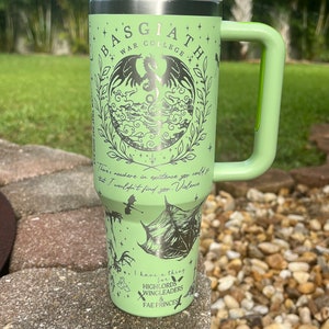 May include: A green stainless steel travel mug with a black and white graphic of a dragon and the text "BASGIATH WAR COLLEGE". The mug has a green handle and a white lid.
