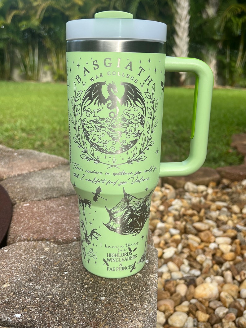 May include: A green stainless steel travel mug with a black and white graphic of a dragon and the text "BASGIATH WAR COLLEGE". The mug has a green handle and a white lid.