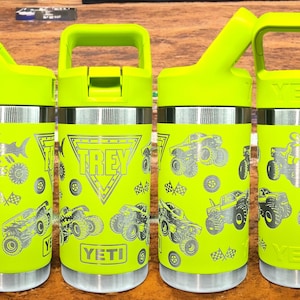 May include: Four neon green insulated water bottles with silver accents. Each bottle features a variety of monster truck graphics, the name "TREY" in a triangle, and the brand "YETI". The bottles have a handle and a flip-top lid.