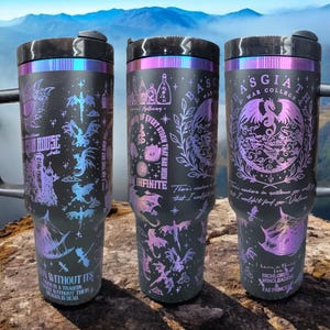 May include: Three black stainless steel travel mugs with a holographic finish. The mugs feature a variety of designs including dragons, a house, and a storm. The text on the mugs reads "Apothecary", "The Eye of Every Storm That Has Been", "Basgiath War College", "I Have a Thing for Highlords, Wingleaders & Fae Princes", "There's no curse in existence you will find that I wouldn't find for you. Valores", "One Without Its Other Is a Tragedy. One Without Their Dragon Is Dead."