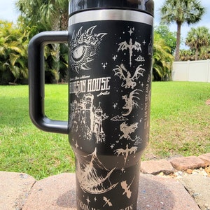 May include: A black stainless steel travel mug with a black handle. The mug features a silver design of a dragon, a castle, and the text "Orson House Arelia" and "I am the sky and the... Dragon without it's rider is a tragedy. A rider without their dragon is dead."