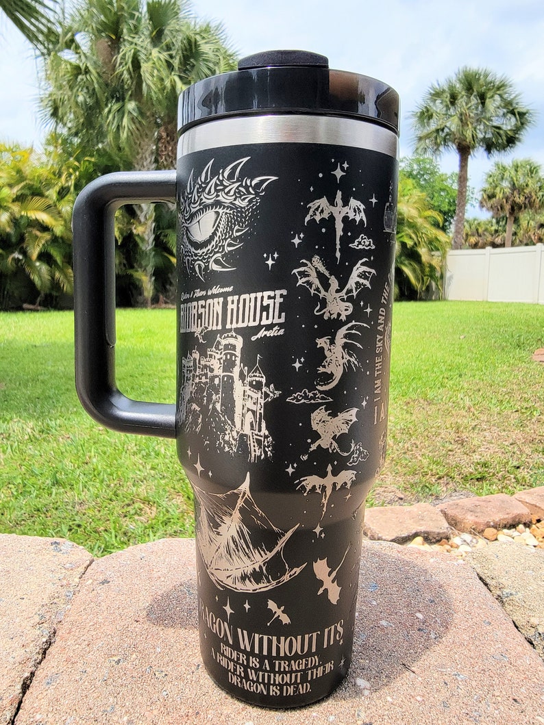 May include: A black stainless steel travel mug with a black handle. The mug features a silver design of a dragon, a castle, and the text "Orson House Arelia" and "I am the sky and the... Dragon without it's rider is a tragedy. A rider without their dragon is dead."