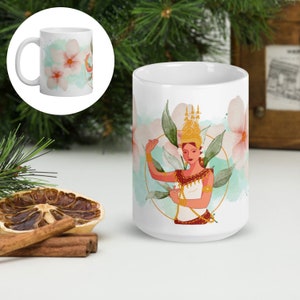 May include: White ceramic mug featuring a colorful illustration of a traditional dancer with a gold headdress and jewelry. The design includes pink flowers and green leaves on a light blue background. The mug has a white handle.