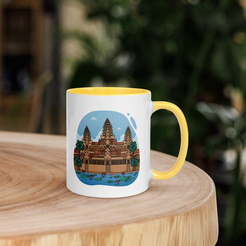 Op de afbeelding: Witte keramische mok met een gele handgreep en binnenkant. De mok heeft een kleurrijke illustratie van een tempel met een blauwe lucht op de achtergrond. De tempel is bruin met groene bomen en een blauwe vijver met waterlelies.