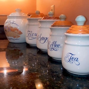 May include: A white ceramic mug with a black handle and the text "But First, Coffee" in a black, handwritten-style font. The mug is on a white surface.