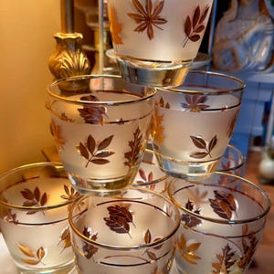 May include: A set of vintage drinking glasses with a frosted finish and gold leaf designs. Each glass has a gold rim and is decorated with various leaf patterns in shades of gold and brown. The glasses are stacked on a white surface.