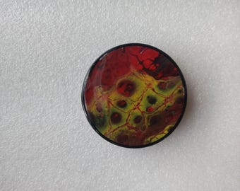 May include: A round, black-rimmed coaster with a colorful abstract design. The design features swirls of red, yellow, and black paint.