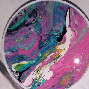 May include: Round, abstract art piece with a white border. The artwork features a vibrant mix of colors, including pink, teal, blue, and black, swirled together with glitter accents. The design has a fluid, marbled appearance, creating a unique decorative item.