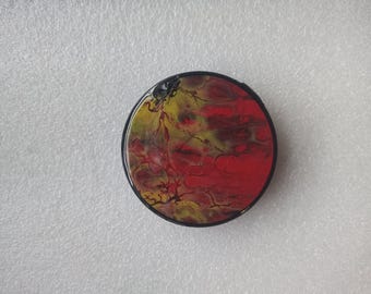 May include: Round, black-rimmed decorative object with a vibrant abstract design. The design features a blend of red, yellow, and black hues, creating a marbled effect. The object is set against a white, textured background.