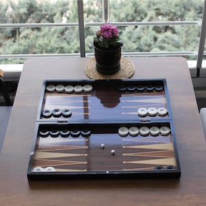 May include: A wooden backgammon board game, open and ready to play, with black and white game pieces. Two dice are on the board. The board is on a brown table, with a potted plant in the background.