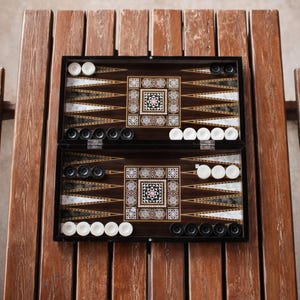 May include: A wooden backgammon set with a dark brown finish, open on a wooden table. The game board features inlaid decorative patterns and triangular points. White and black game pieces are arranged on the board, ready for play.