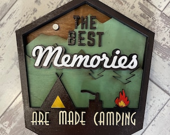 May include: A wooden sign with a brown frame and a green background. The sign says "The Best Memories Are Made Camping" with a stylized tent, campfire, and mountains in the background.