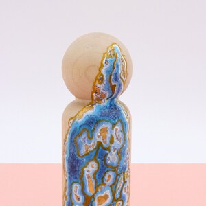 Op de afbeelding: Een houten pop geschilderd met een blauw en gouden agaatpatroon. De pop staat op een roze oppervlak. @littleskweeks
