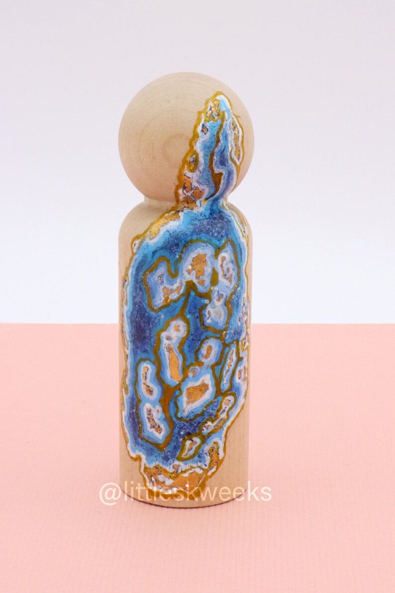 Op de afbeelding: Een houten pop geschilderd met een blauw en gouden agaatpatroon. De pop staat op een roze oppervlak. @littleskweeks