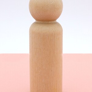 Op de afbeelding: Een houten pop met een rond hoofd en een cilindrisch lichaam. De pop is ongeverfd en heeft een natuurlijke houten afwerking.