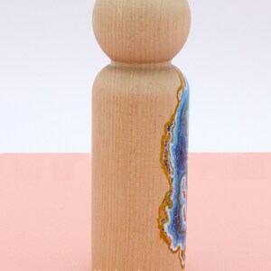 Op de afbeelding: Een houten pop met een geschilderd agaatontwerp aan de ene kant. Het agaatontwerp heeft blauwe, witte en gouden kleuren.