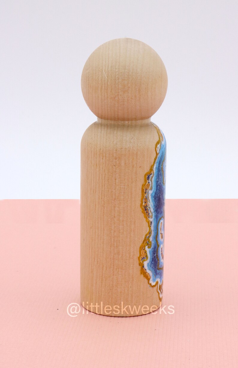 Op de afbeelding: Een houten pop met een geschilderd agaatontwerp aan de ene kant. Het agaatontwerp heeft blauwe, witte en gouden kleuren.