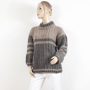 Könnte beinhalten: Ein gemütlicher Pullover in neutralen Tönen mit hohem Kragen und Rippenstruktur. Das Design weist ein Colorblocking-Muster mit Grau- und Beigetönen auf. Die Ärmel sind dunkelgrau, der Körper hat einen horizontalen Streifen.