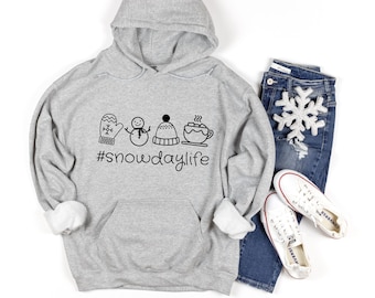 May include: A heather gray hoodie with a black graphic of winter icons, including a mitten, snowman, hat, and hot cocoa, with the text "#snowdaylife." The hoodie is paired with blue jeans, white sneakers, and a snowflake decoration.